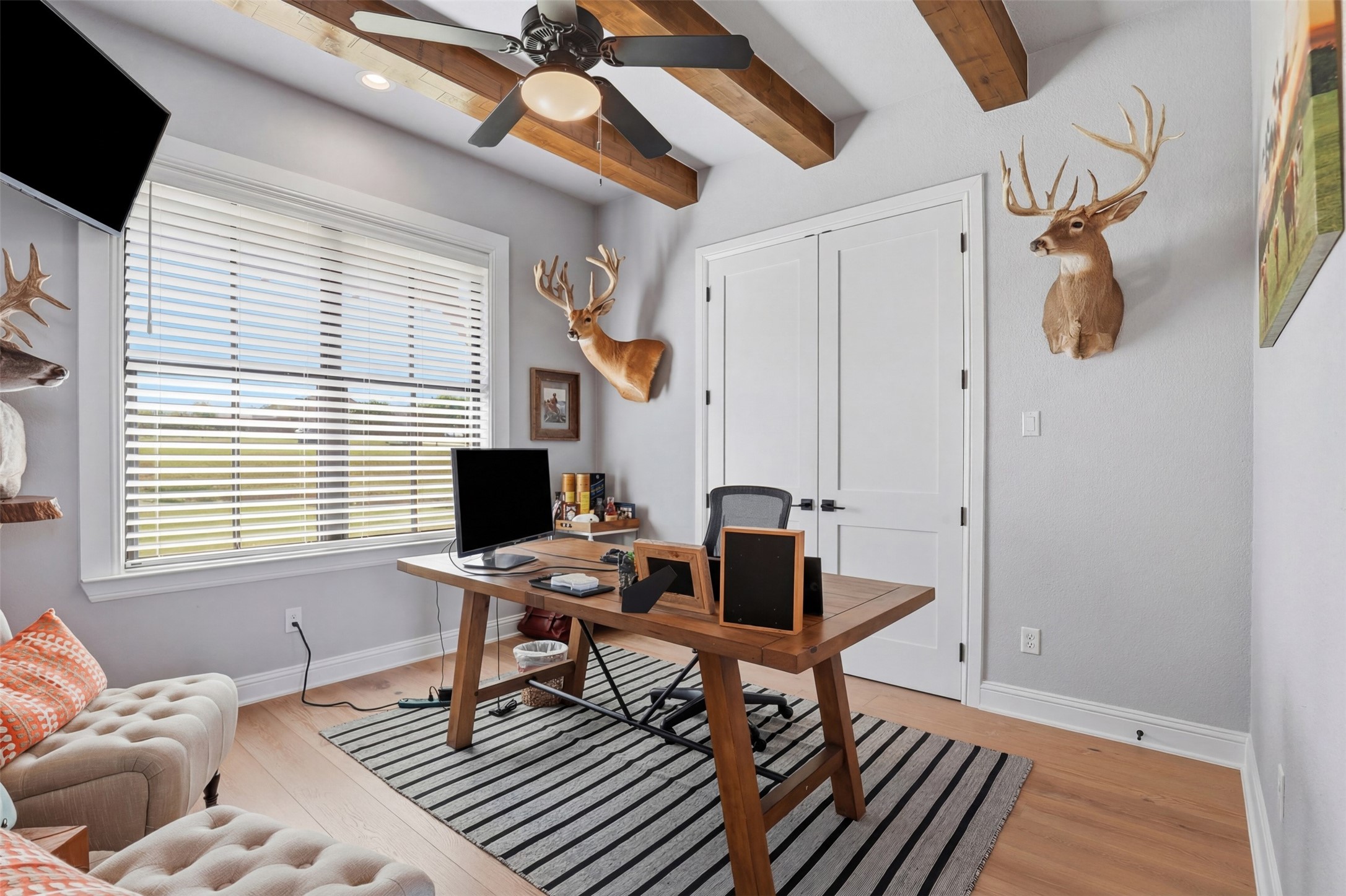 Furnished home office with a wooden desk beneath exposed beams and mounted deer heads on the walls.