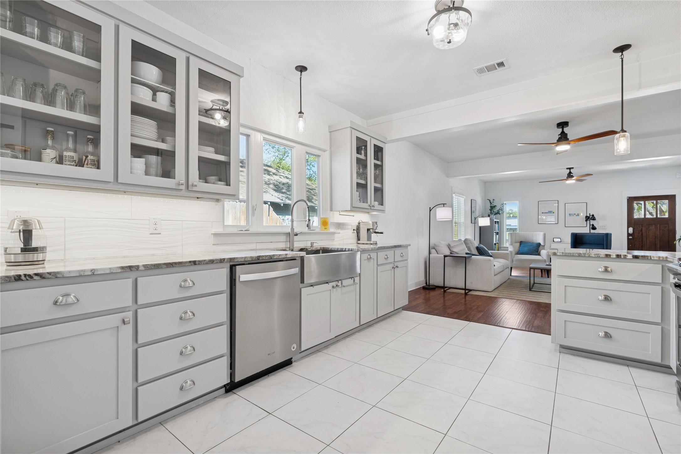 Bright white kitchen with glass-front cabinets and farmhouse sink, opening to a furnished living room.