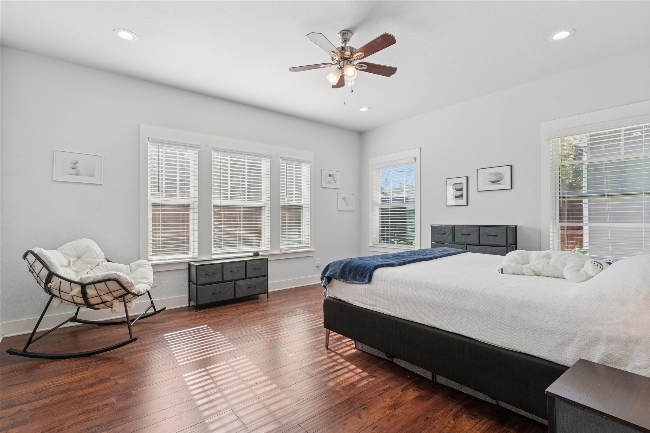 Bright bedroom with a large bed, dark wood floors, and a cushioned rocking chair by the windows.