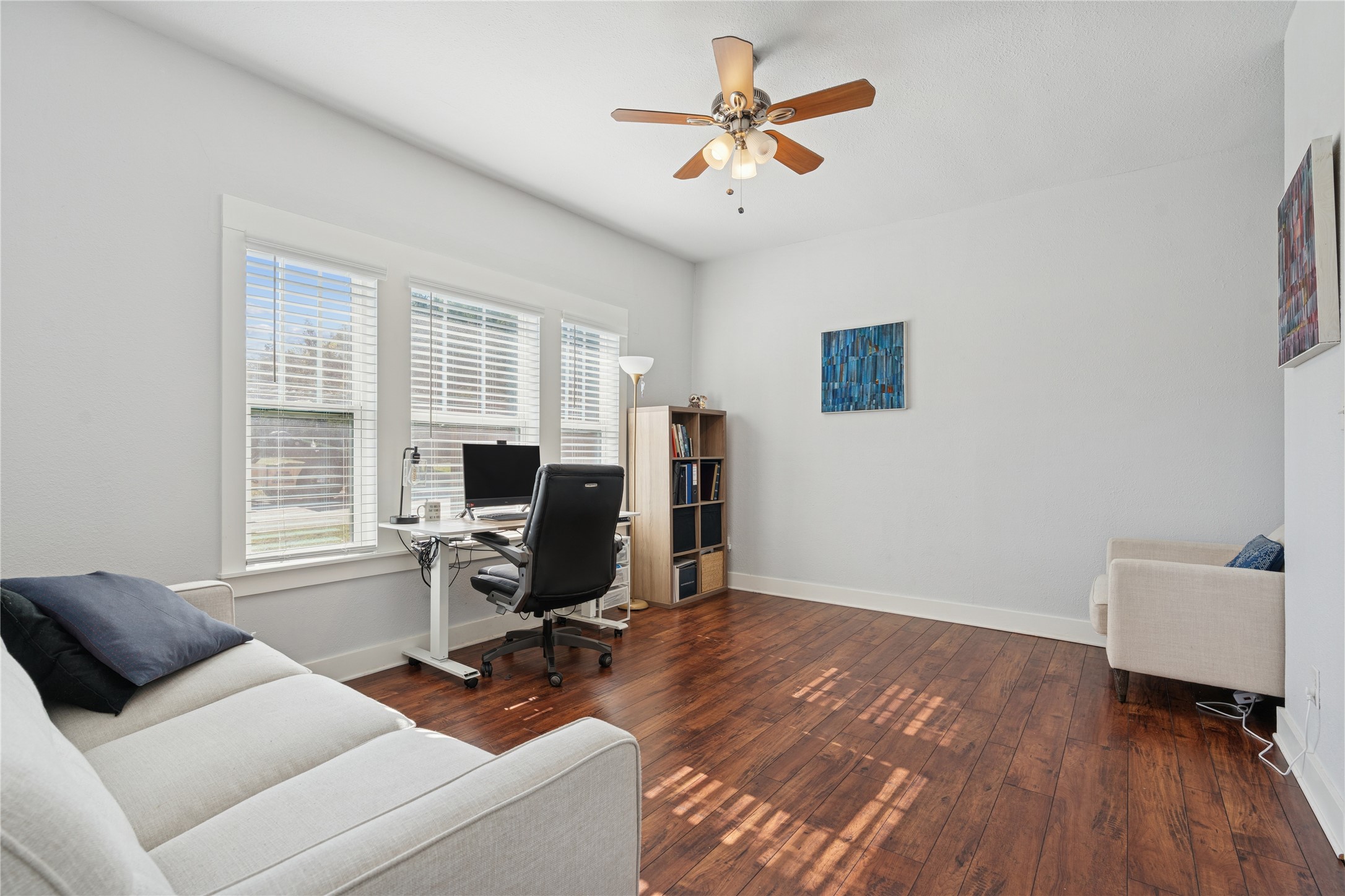 Home office with a desk by three windows, hardwood floors, and light-colored sofas.