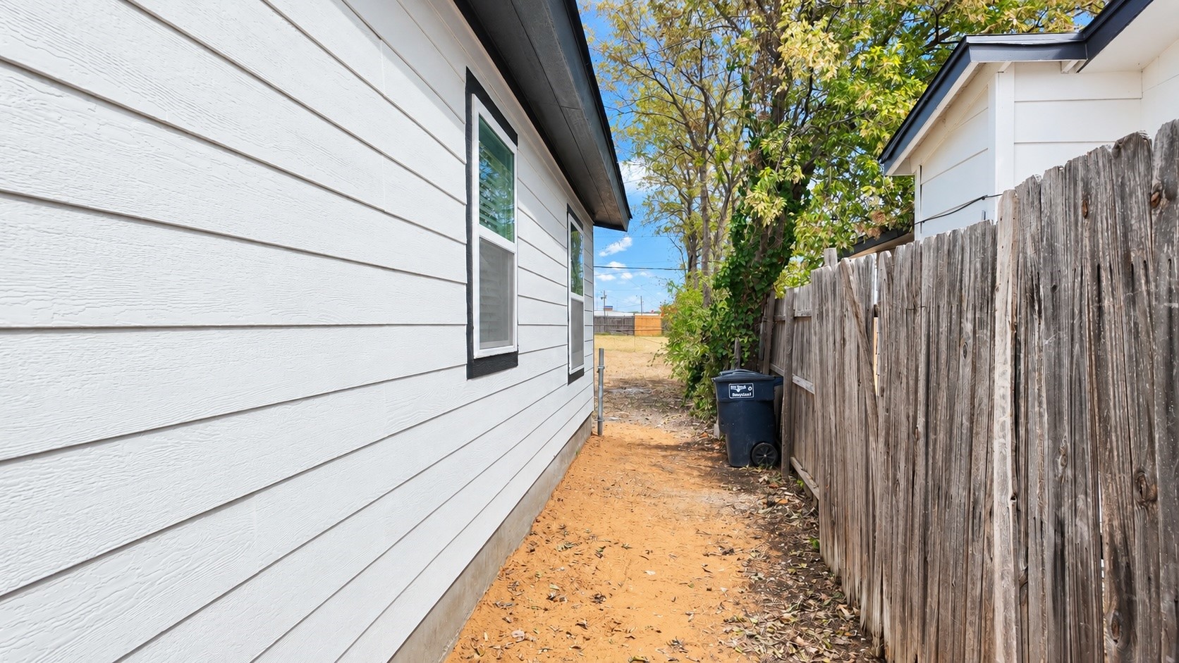 Narrow sandy side yard between a white-sided house and a weathered wooden fence.