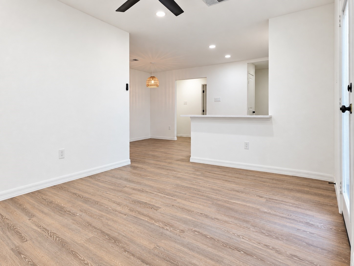 Empty open-plan living and dining area with wood-look floors, a pass-through counter, and glazed doors.