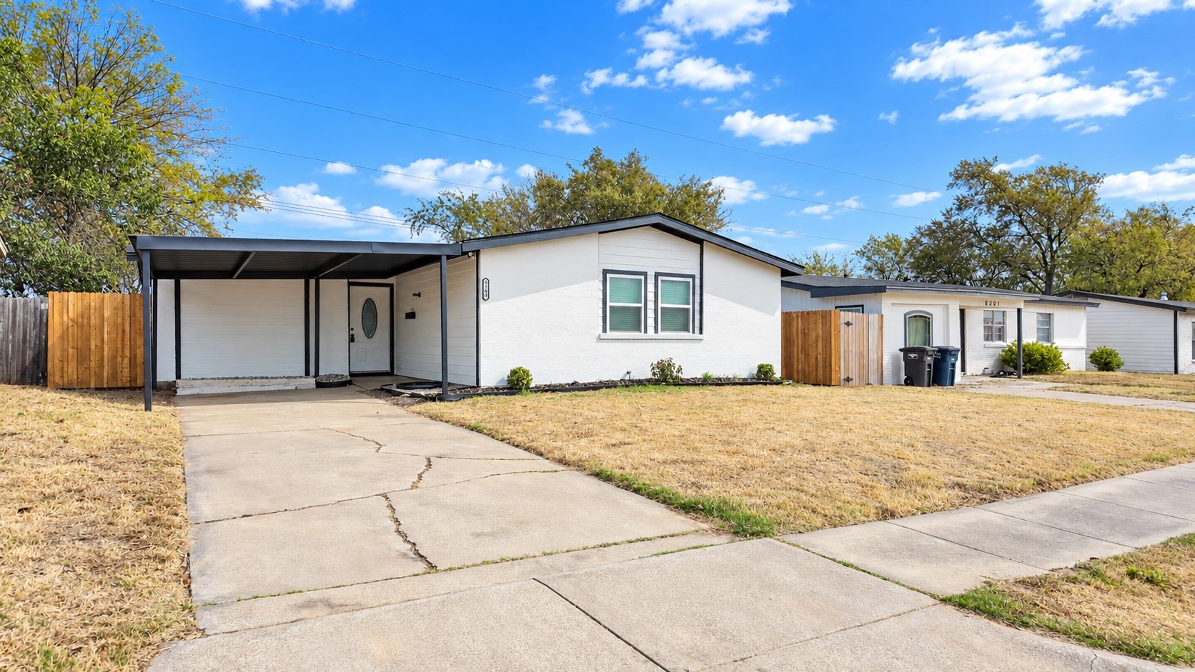 White single-story home with black trim, covered carport, concrete driveway, and wooden fence.
