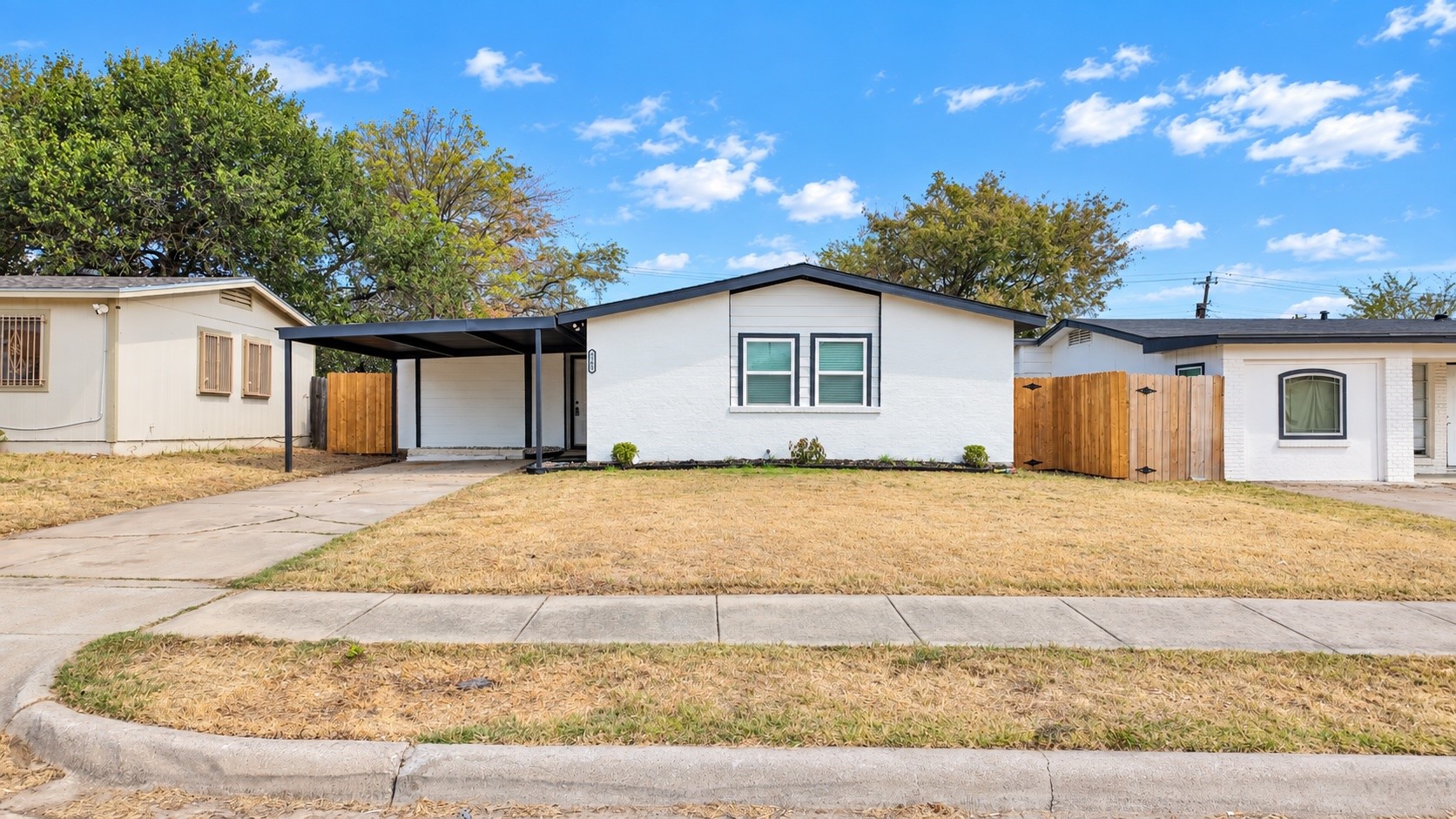 White single-story house with a covered carport, wooden fences, and a dry front lawn.