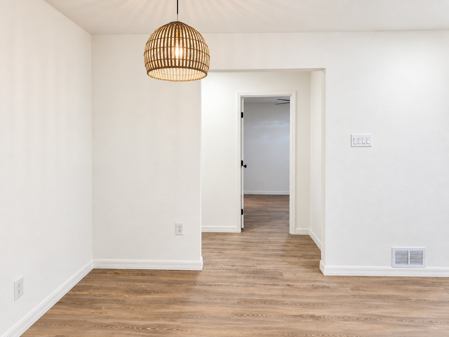Empty room with wood-look flooring and a woven pendant light, looking through an open doorway.