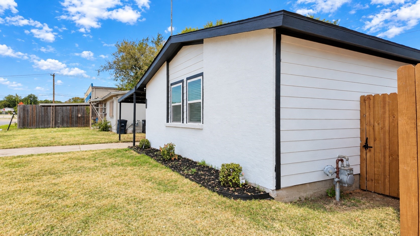 White-sided home with black trim, landscaped foundation, and wooden gate beside the yard.