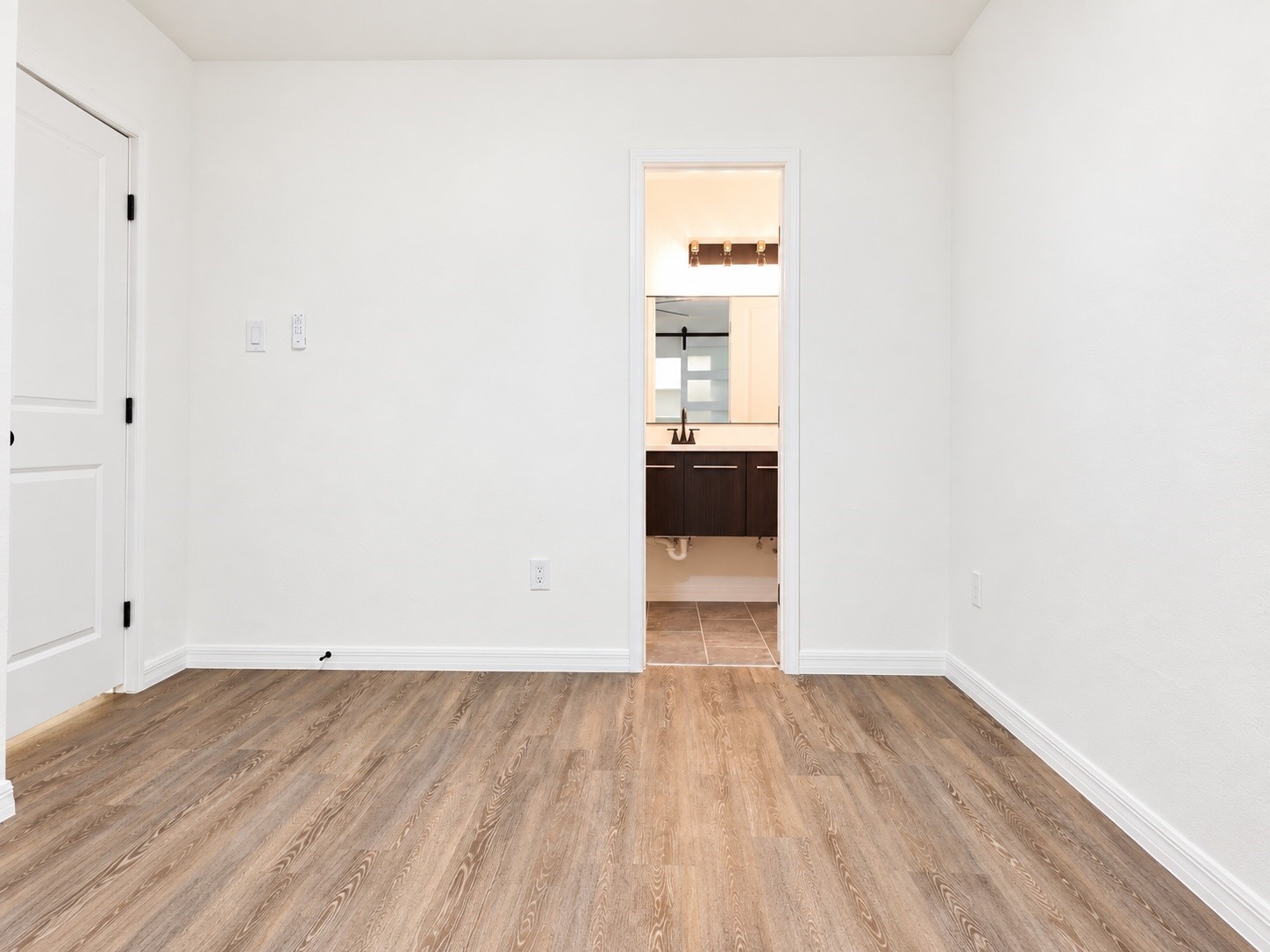 Empty room with wood-look flooring and a doorway into a bathroom with a dark vanity.
