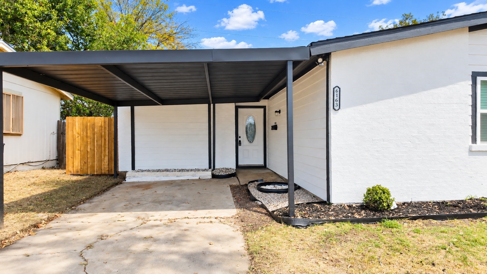 White house with a black covered carport, concrete driveway, and wooden side fence.