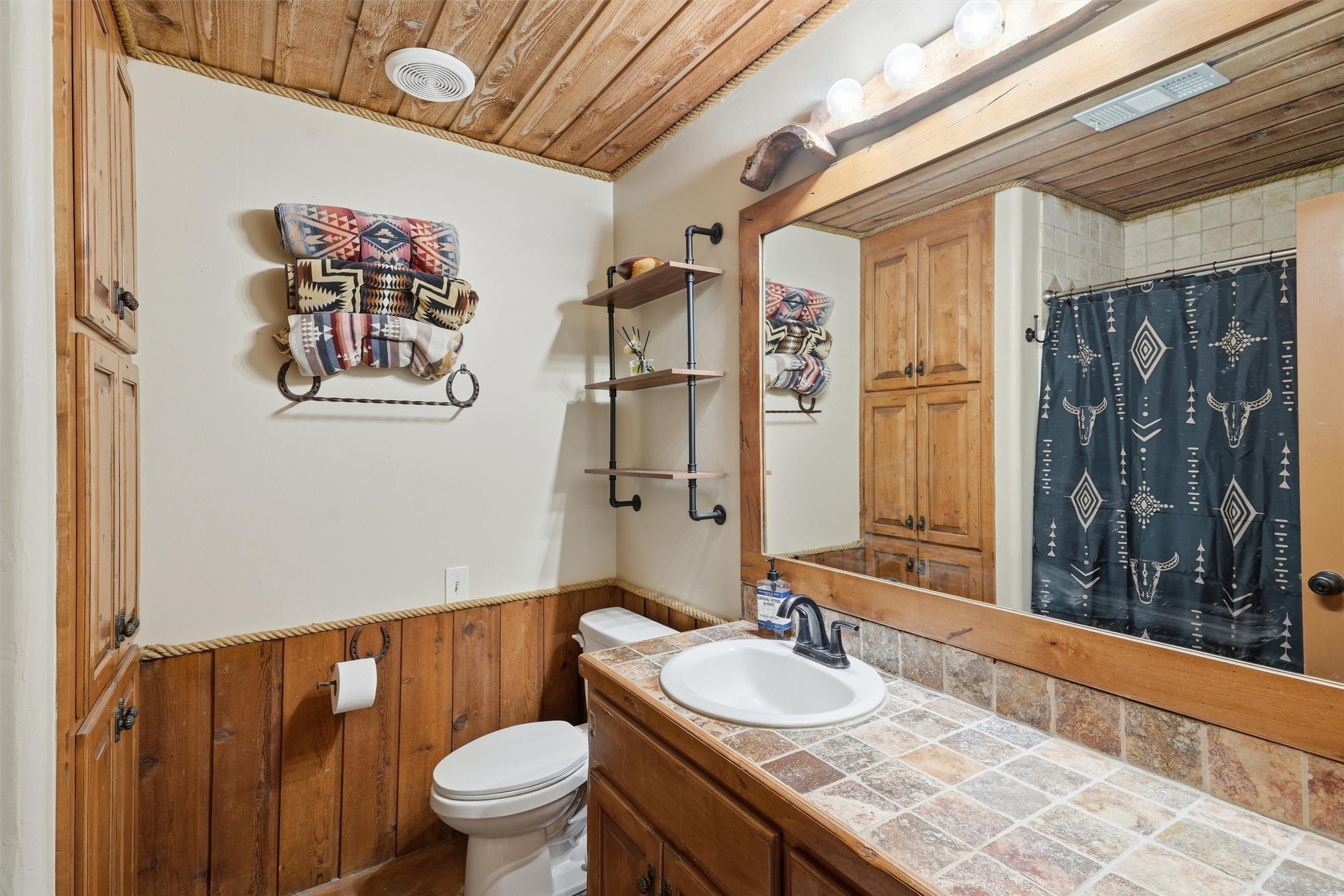 Rustic bathroom with a tiled vanity, wood cabinetry, exposed wood ceiling, and patterned shower curtain.