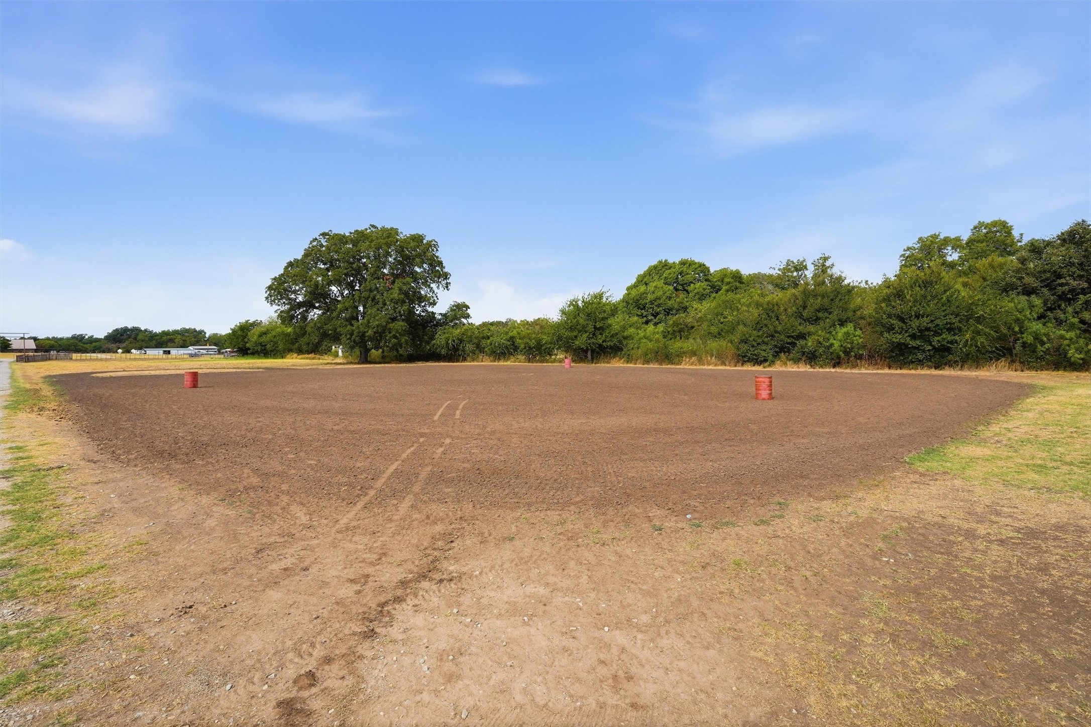 Dirt riding arena with three red barrels, grassy edges, and trees along the far side.