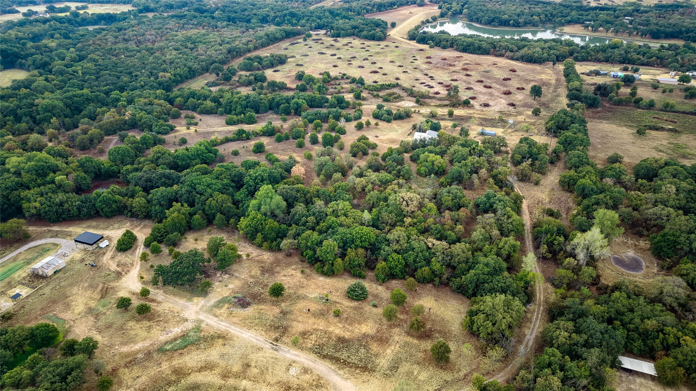 Aerial view of wooded acreage with open clearings, dirt tracks, outbuildings, and a pond.