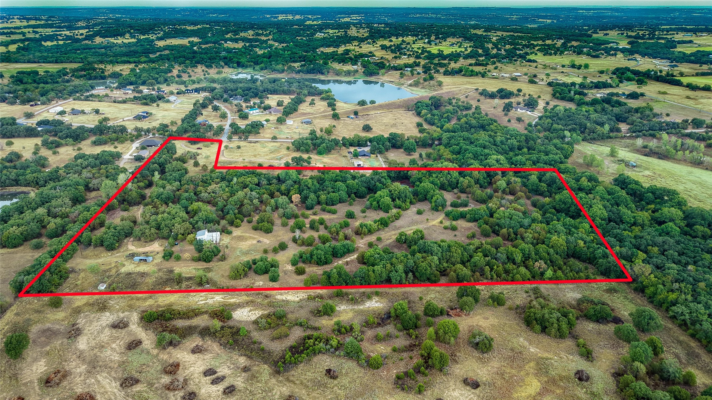 Aerial view of wooded acreage outlined in red, with a pond and neighboring properties beyond.