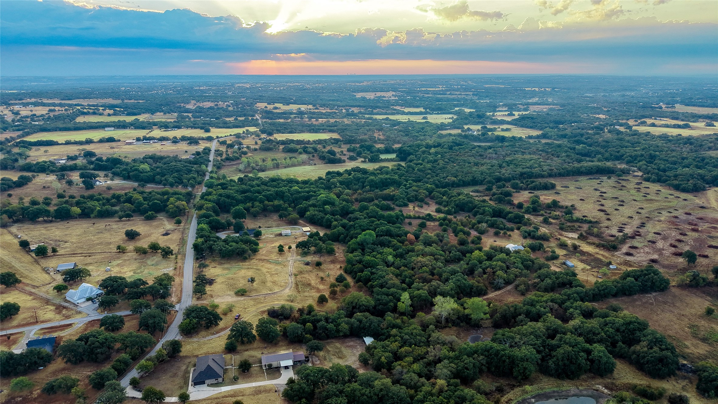 Aerial view of rural acreage with wooded fields, scattered homes, and a small pond at sunset.