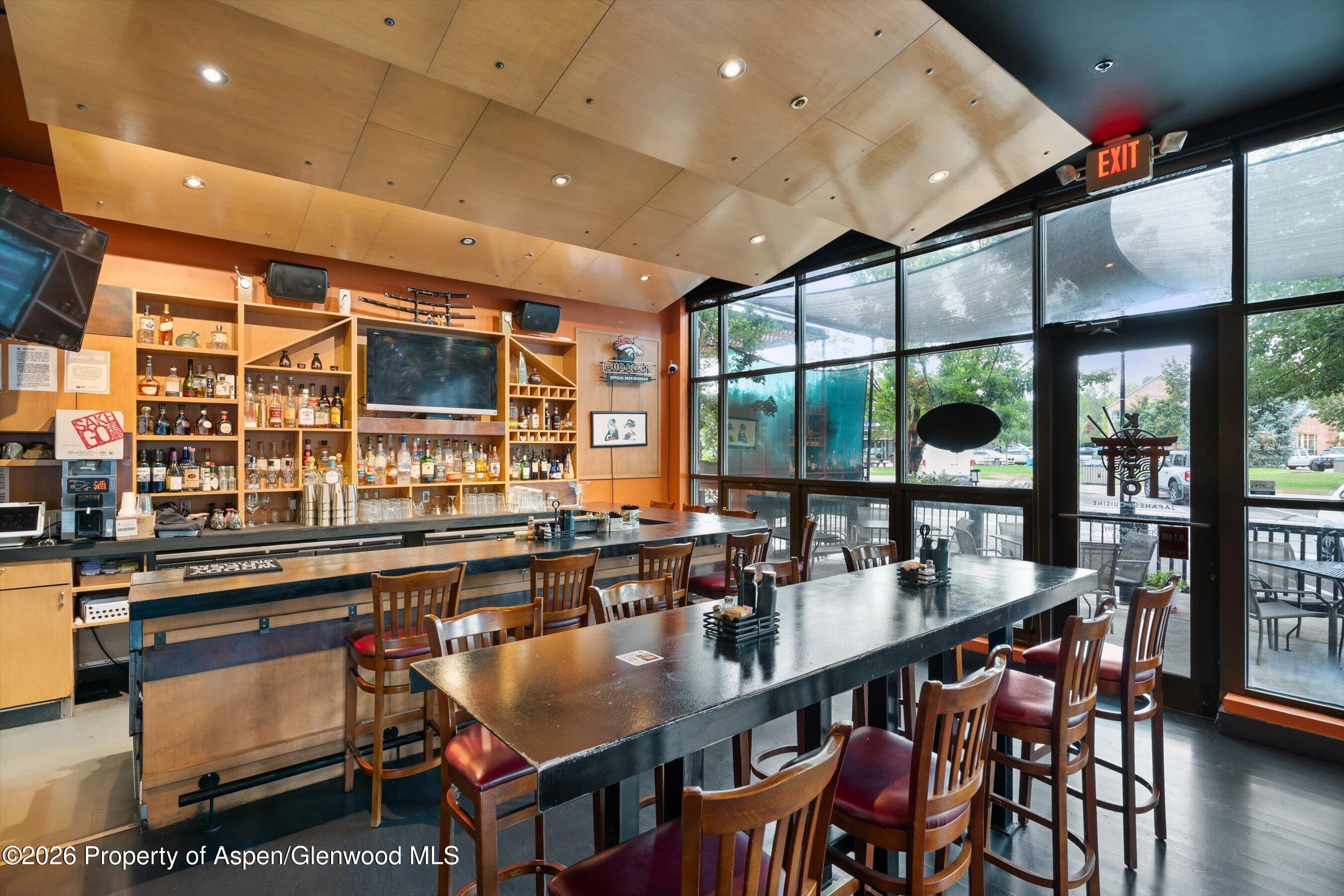 Restaurant bar with liquor shelves, wooden dining tables, and tall street-facing windows.
