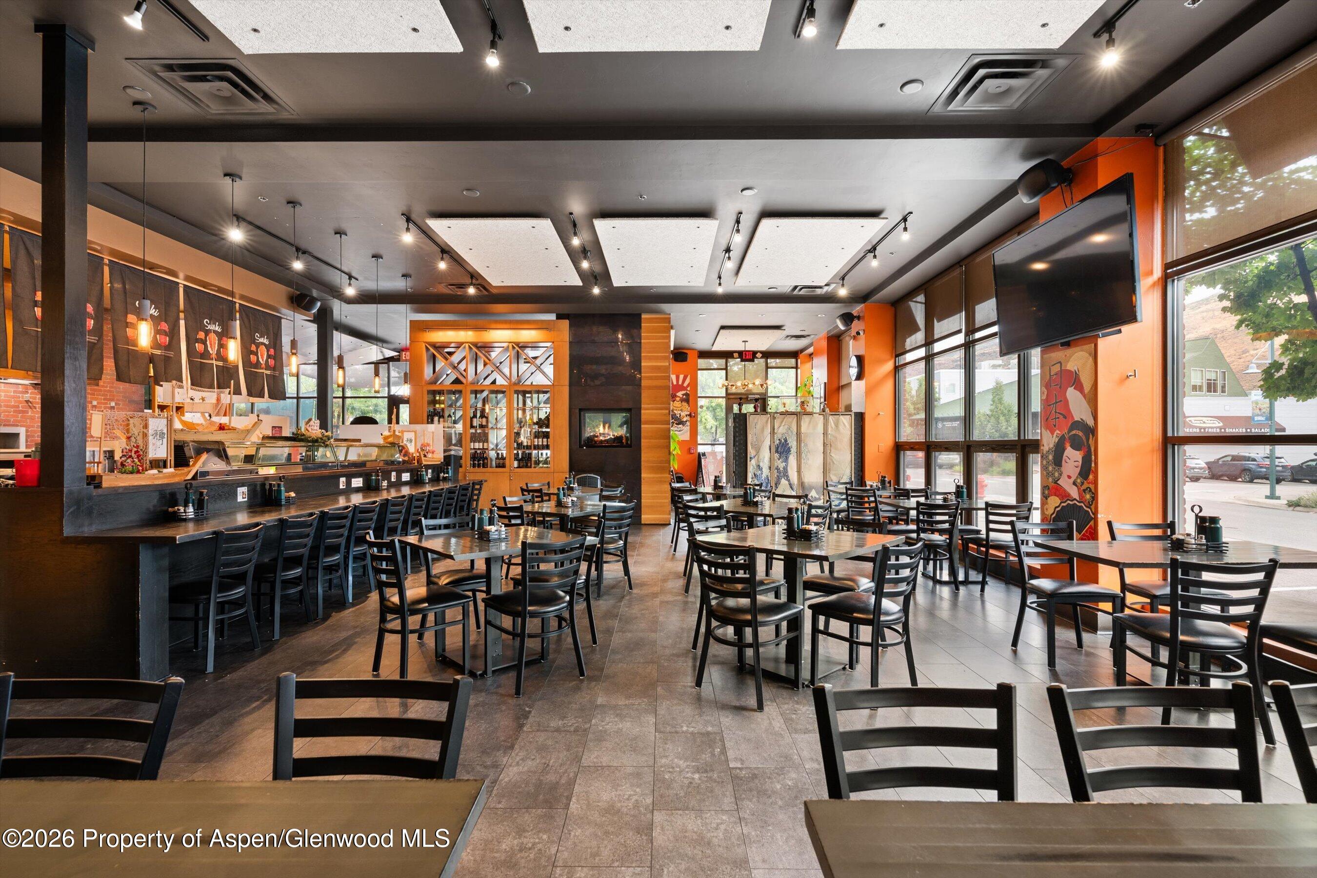 Restaurant dining room with a sushi counter, black tables and chairs, orange walls, and large windows.