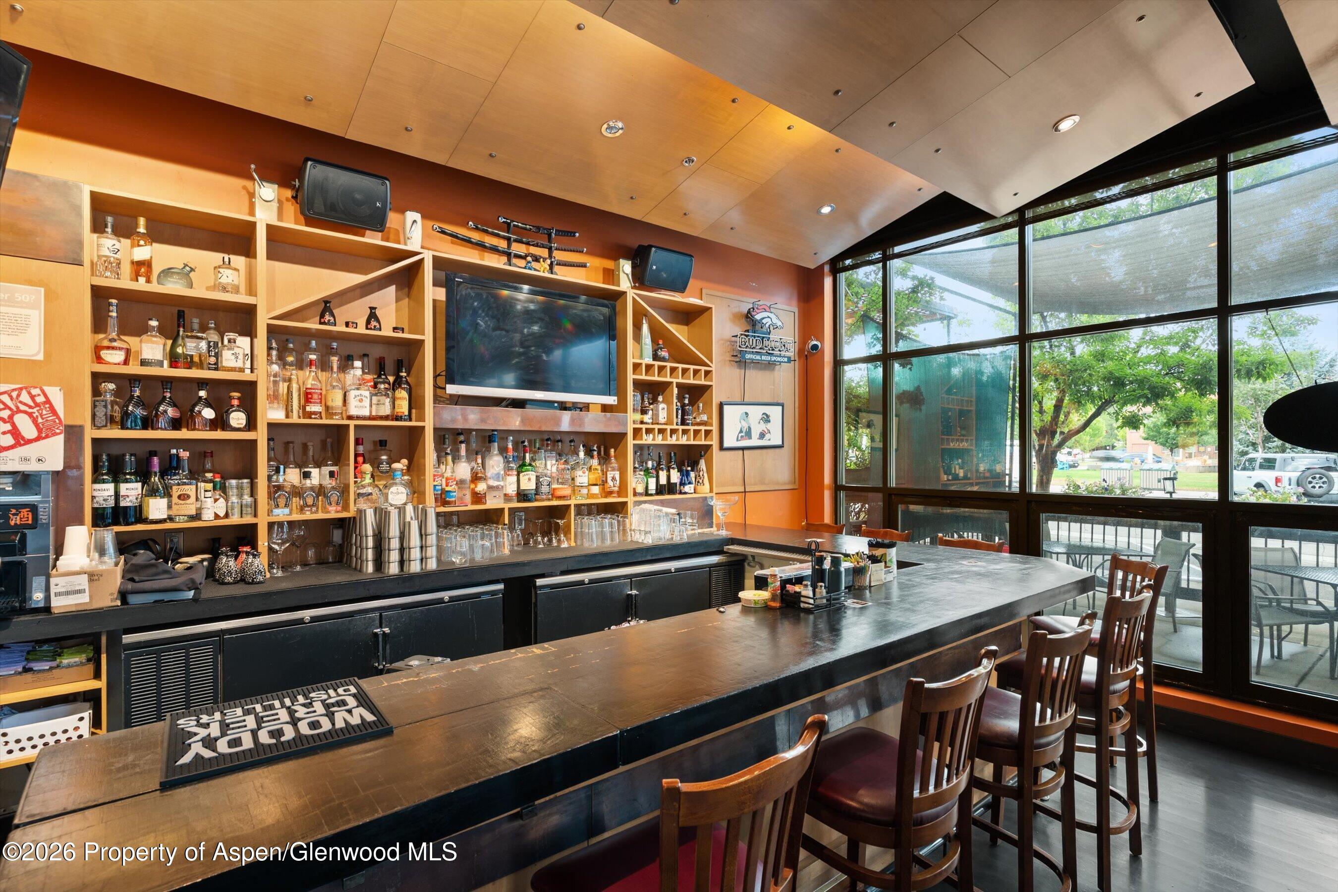 Bar interior with liquor shelves, a long counter, wooden stools, and large windows facing a patio.