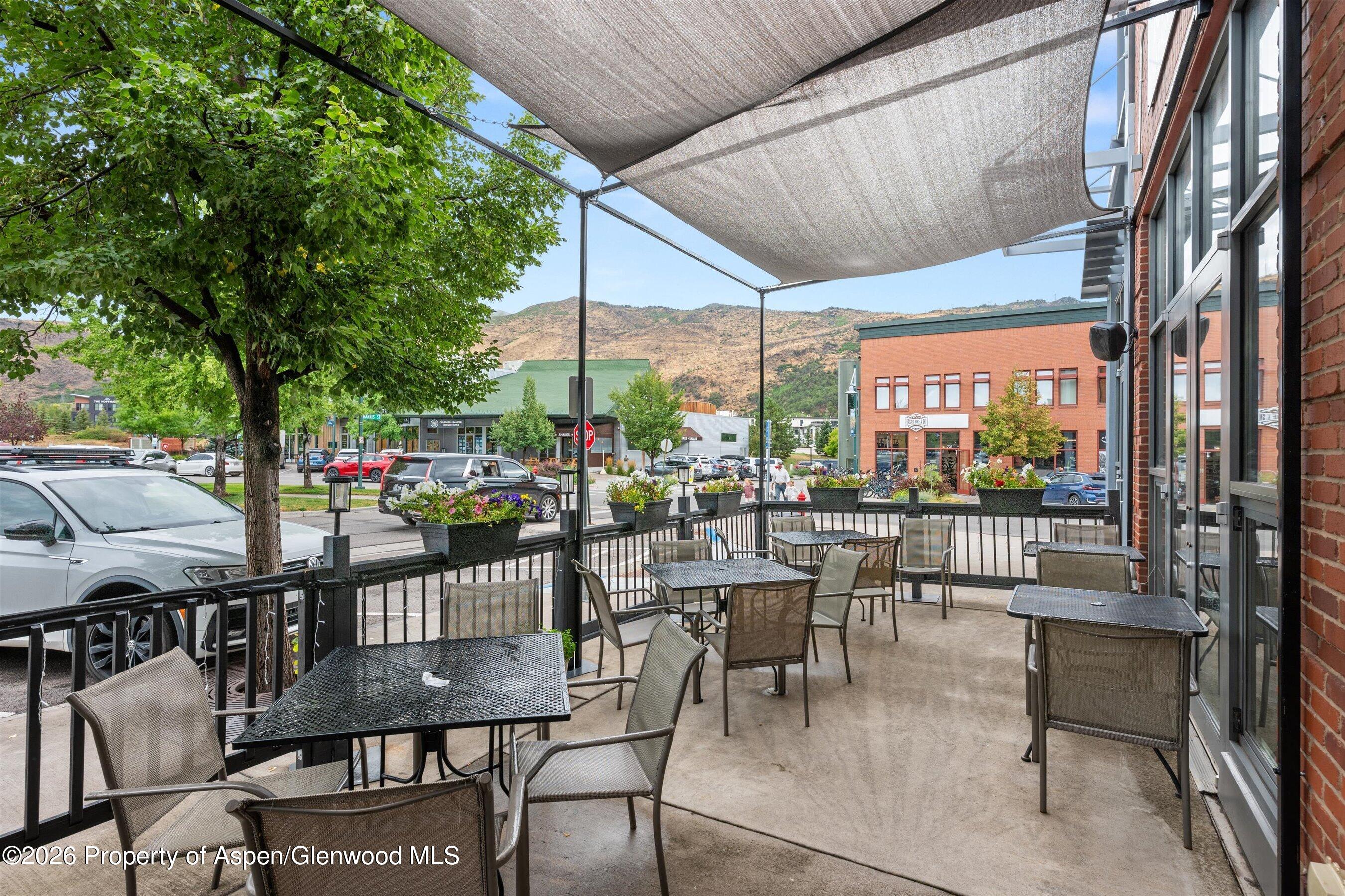 Outdoor patio with café seating beneath shade sails, overlooking a tree-lined street and mountain hills.