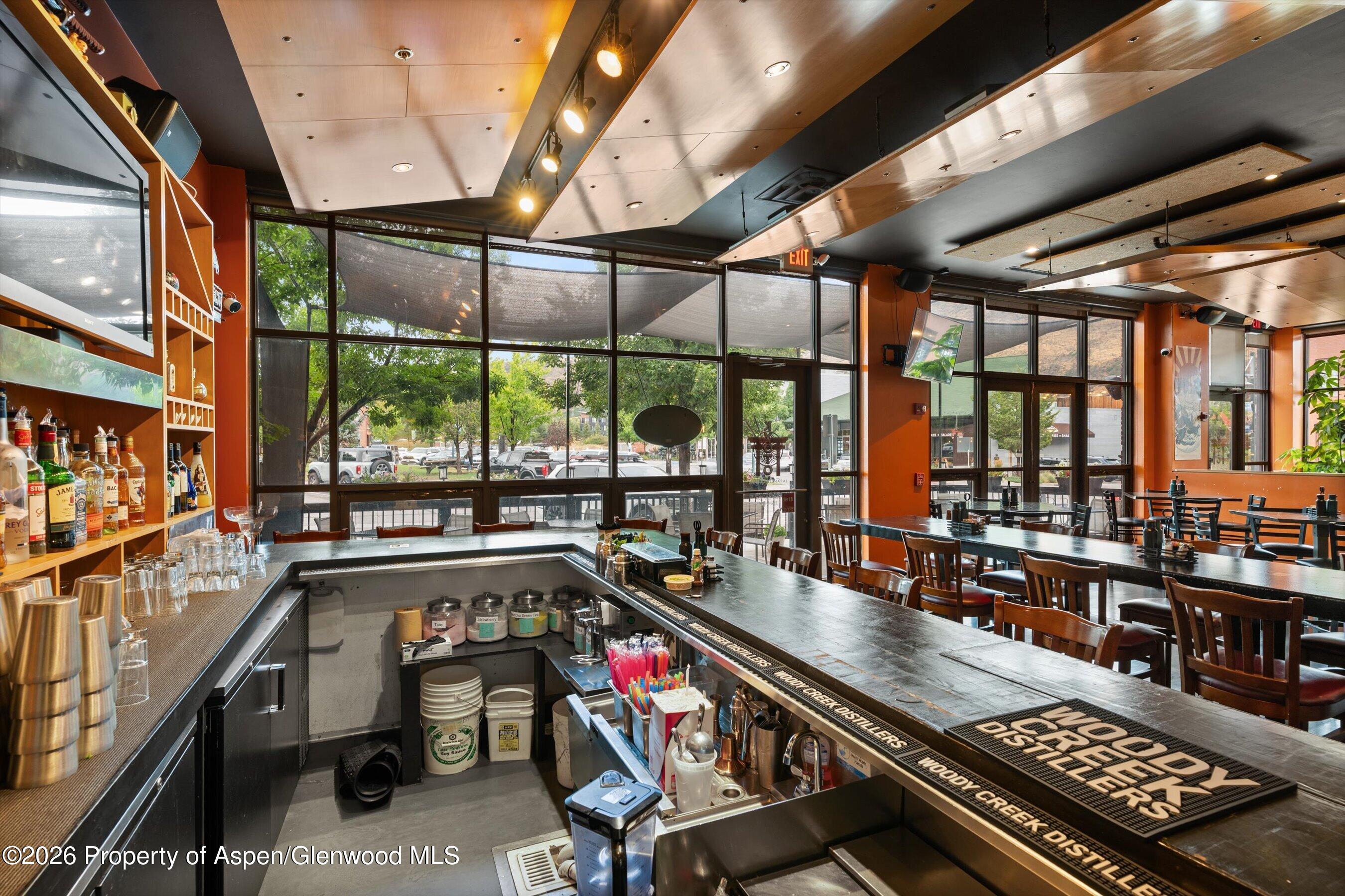 Restaurant bar and dining room with liquor shelves, wooden tables, and tall street-facing windows.