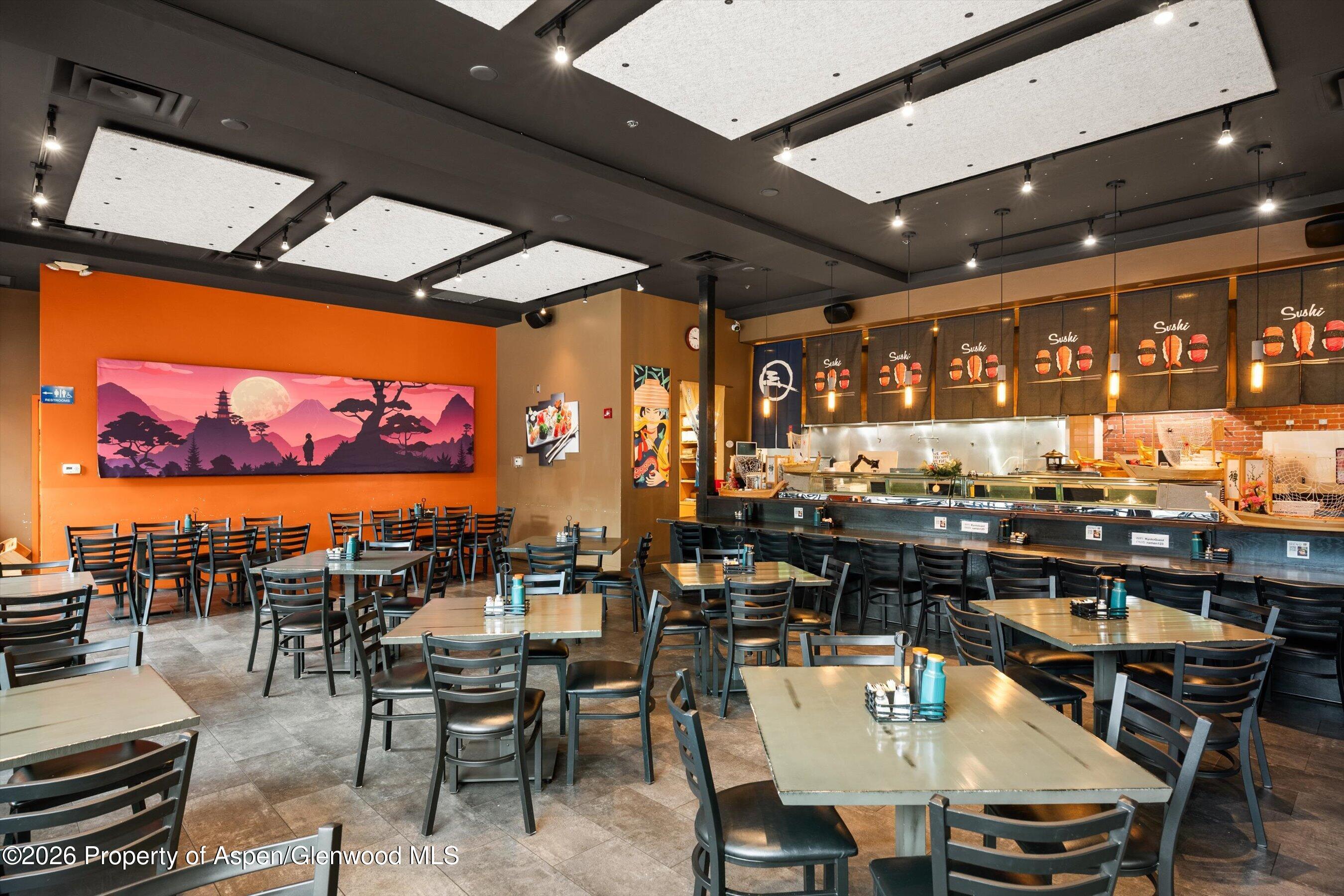 Sushi restaurant dining room with a long counter, rows of tables, and a large mural on an orange wall.