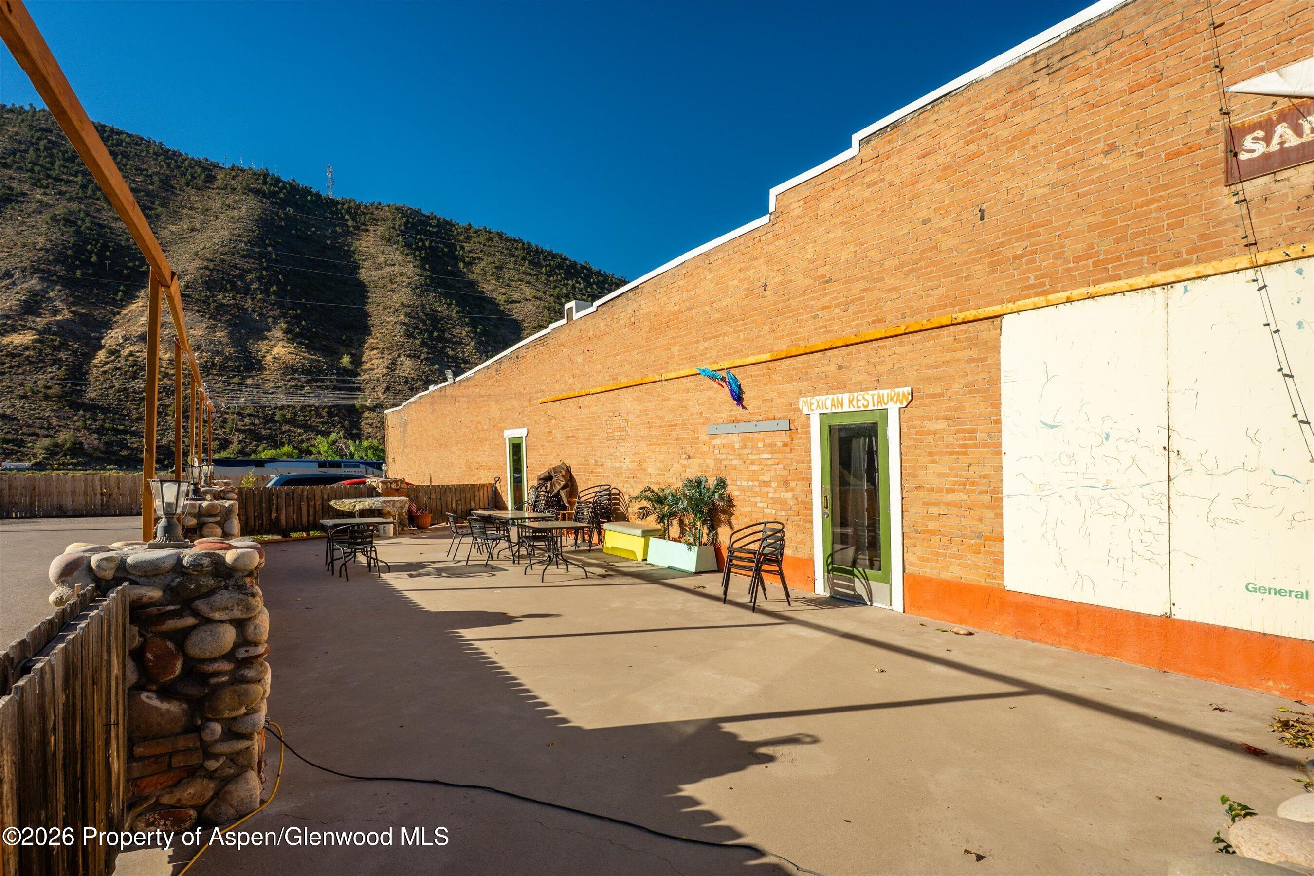 Brick commercial building with a paved outdoor dining patio, metal tables, and a mountain hillside behind it.