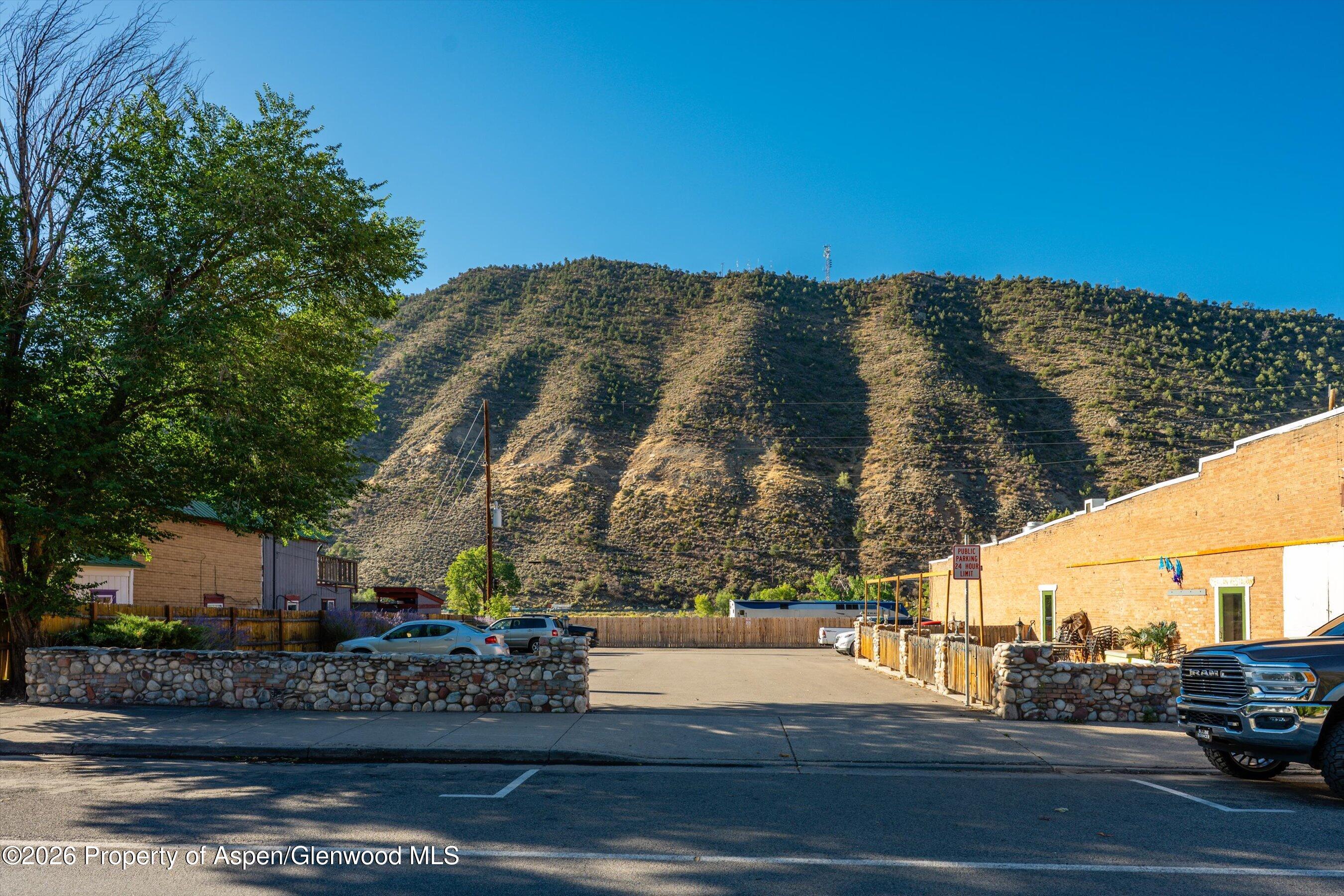 Paved commercial lot between brick buildings, with a wooded hillside behind it.