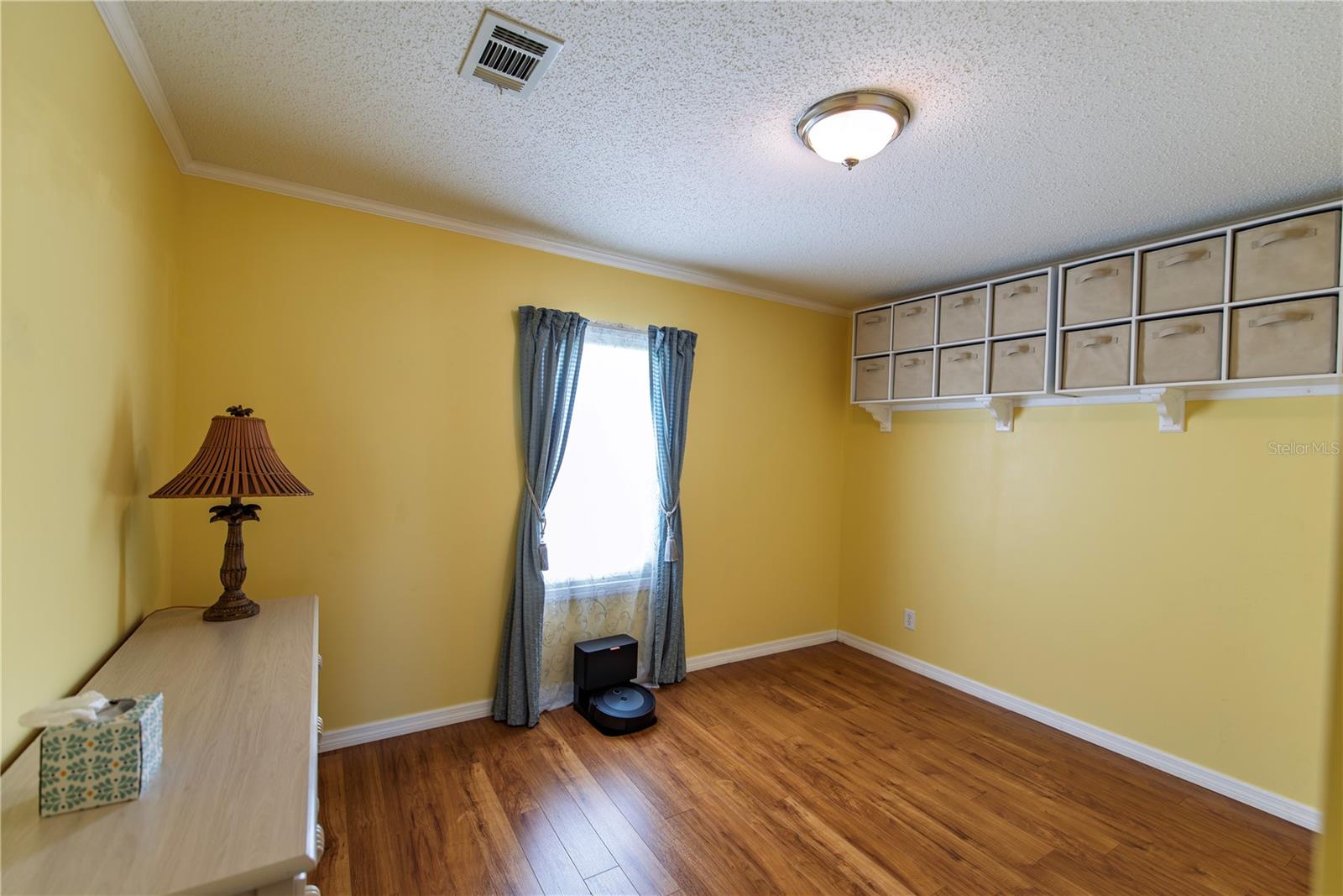 Yellow-walled room with wood flooring, a desk, curtained window, and wall-mounted storage cubbies.