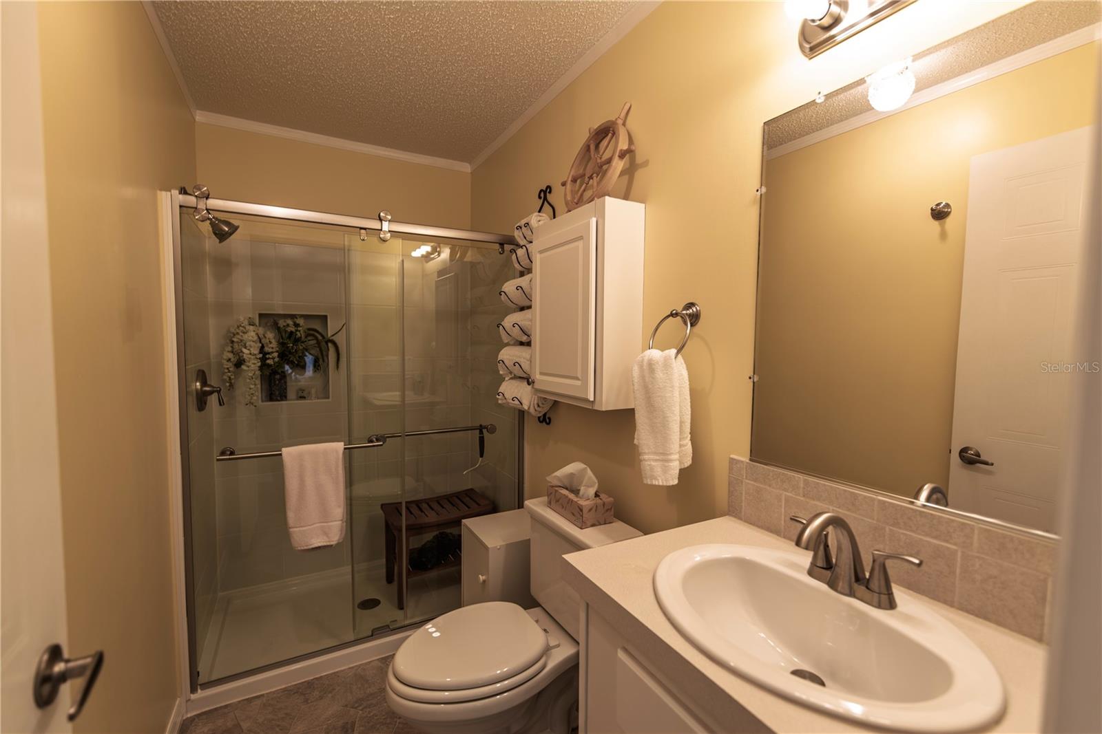 Bathroom with a glass shower, white vanity, large mirror, and nautical ship's wheel decor.