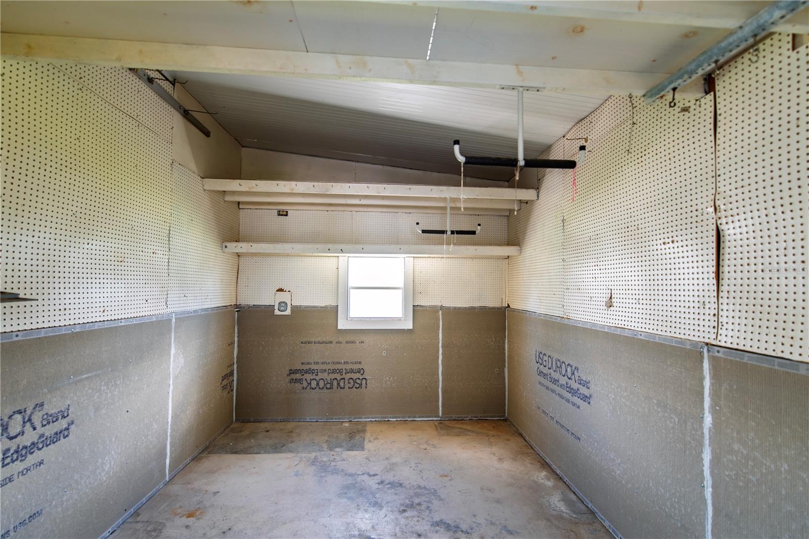 Empty unfinished garage with pegboard walls, overhead shelves, and a small rear window.