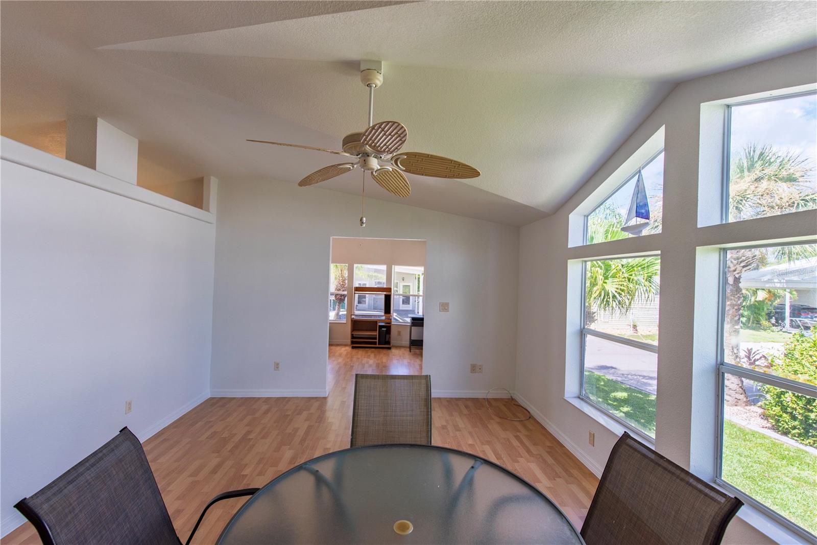 Dining area with vaulted ceiling, large windows, and a glass-top table overlooking a palm-lined yard.