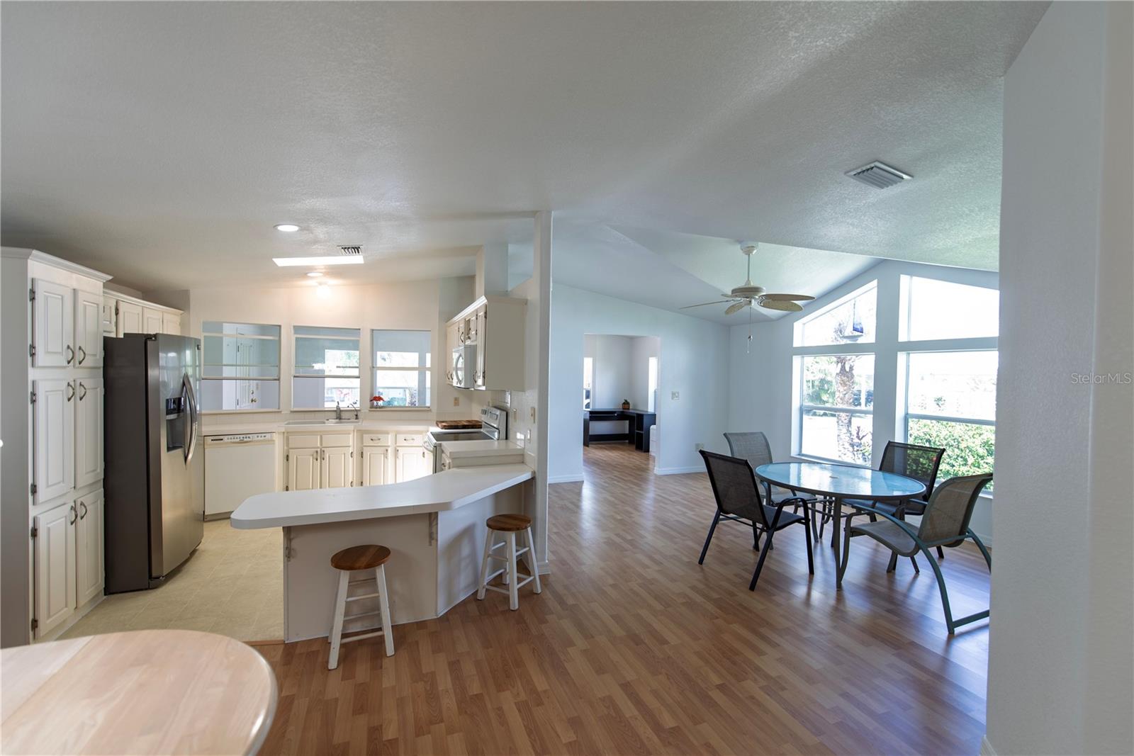 Open kitchen with white cabinets and curved breakfast bar beside a bright dining area with large windows.