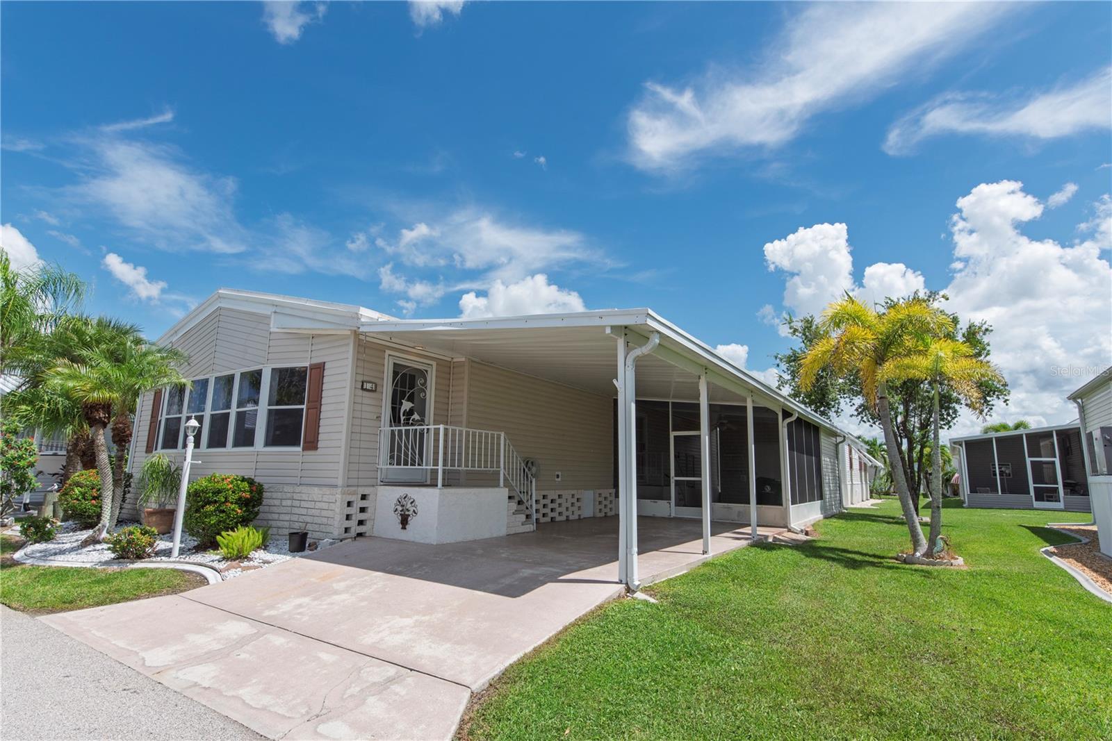 Manufactured home with a covered carport, screened porch, palm trees, and a landscaped lawn.