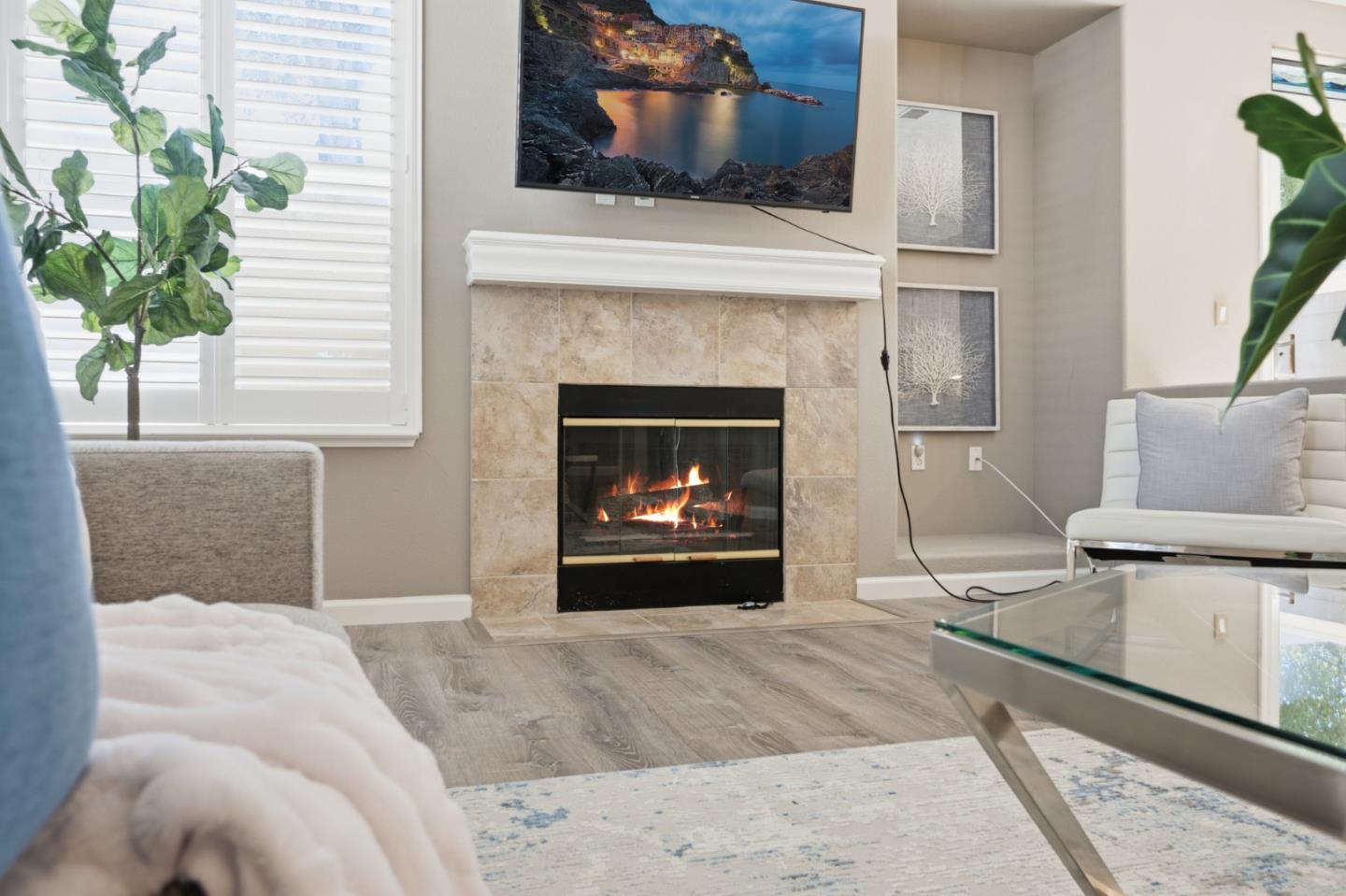 Living room with a lit tiled fireplace, wall-mounted television, and white plantation shutters.