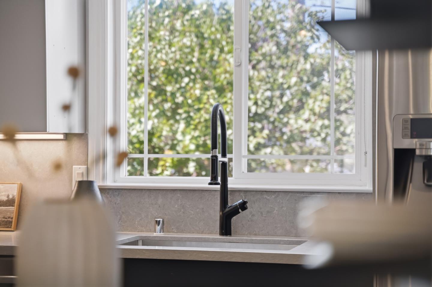 Kitchen sink with black pull-down faucet beneath a large window overlooking leafy greenery.