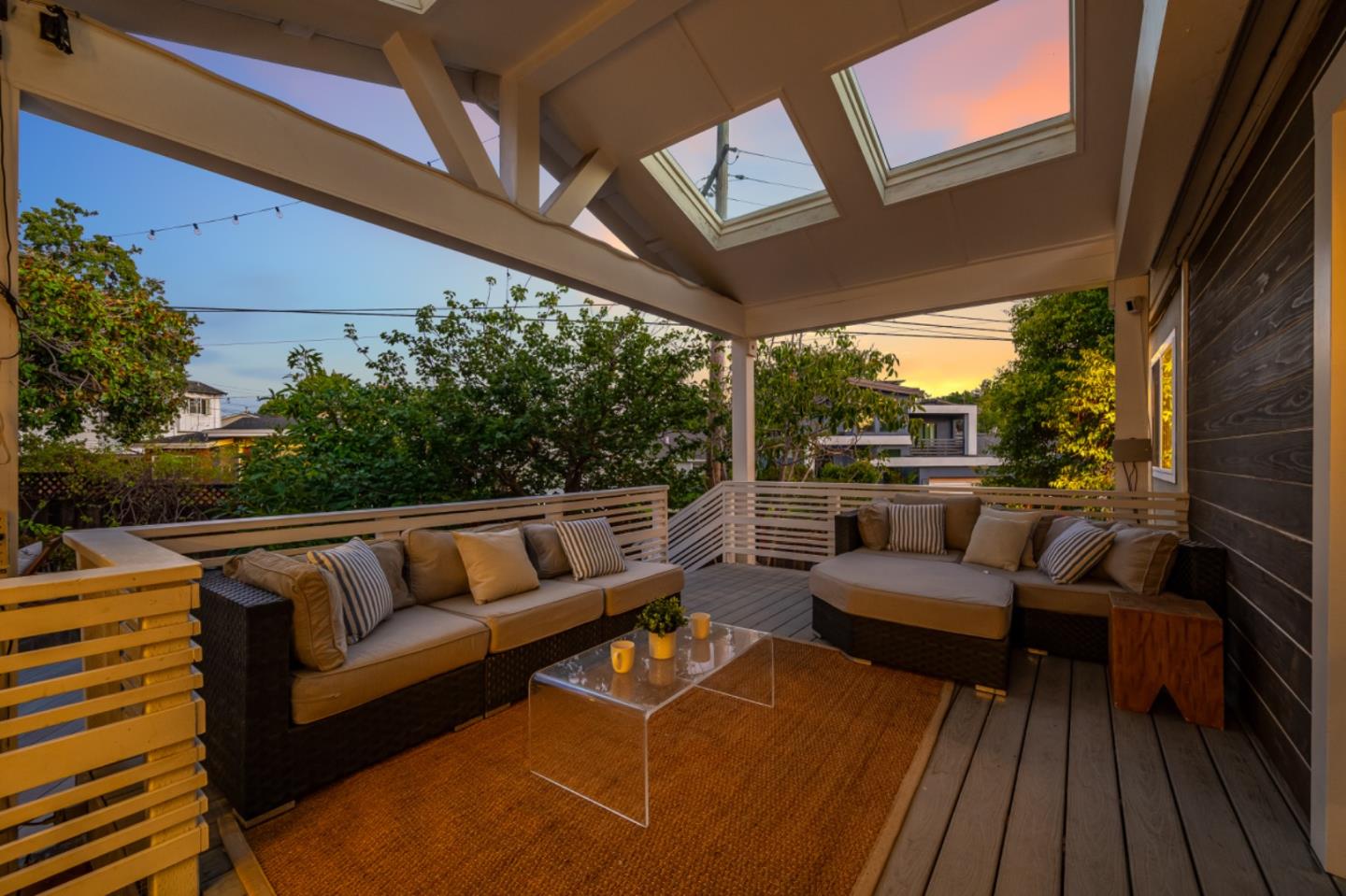 Covered deck with sectional seating, roof skylights, and trees beyond at sunset.