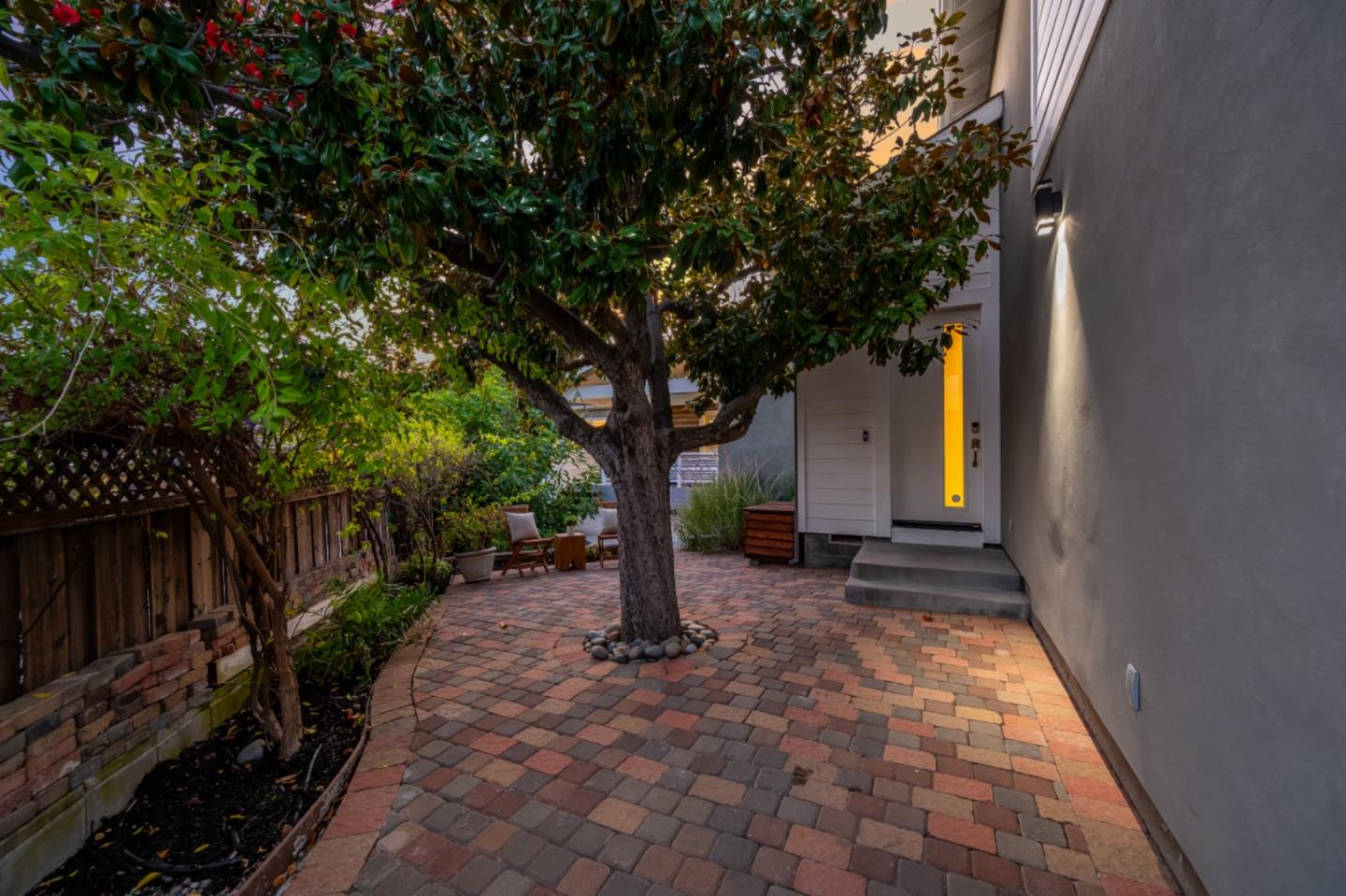 Front courtyard with multicolored pavers, a mature tree, patio seating, and a modern entry door.