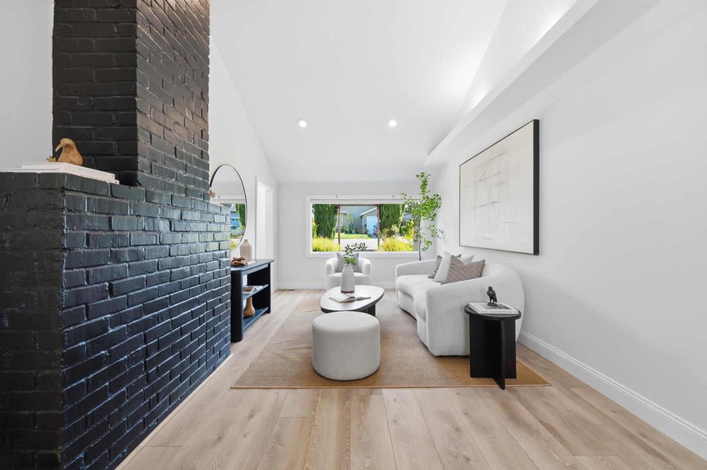 Staged living room with a black brick fireplace, white sofa, and garden views through a wide window.