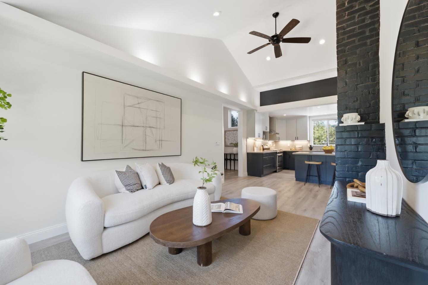 Living room with a vaulted ceiling, white sofa, dark brick fireplace, and open kitchen.