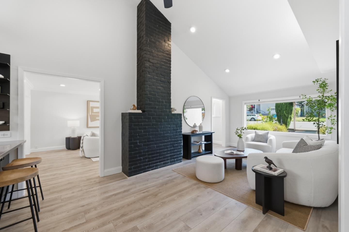 Staged living room with a tall black brick fireplace, vaulted ceiling, and white seating by a large window.