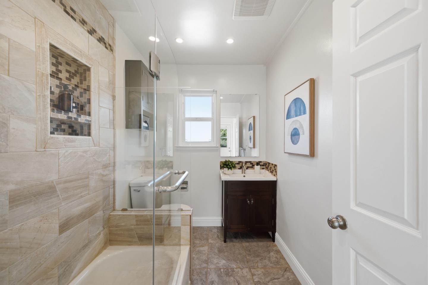 Bathroom with glass-enclosed tub, beige stone tile, mosaic shower niche, and dark wood vanity.