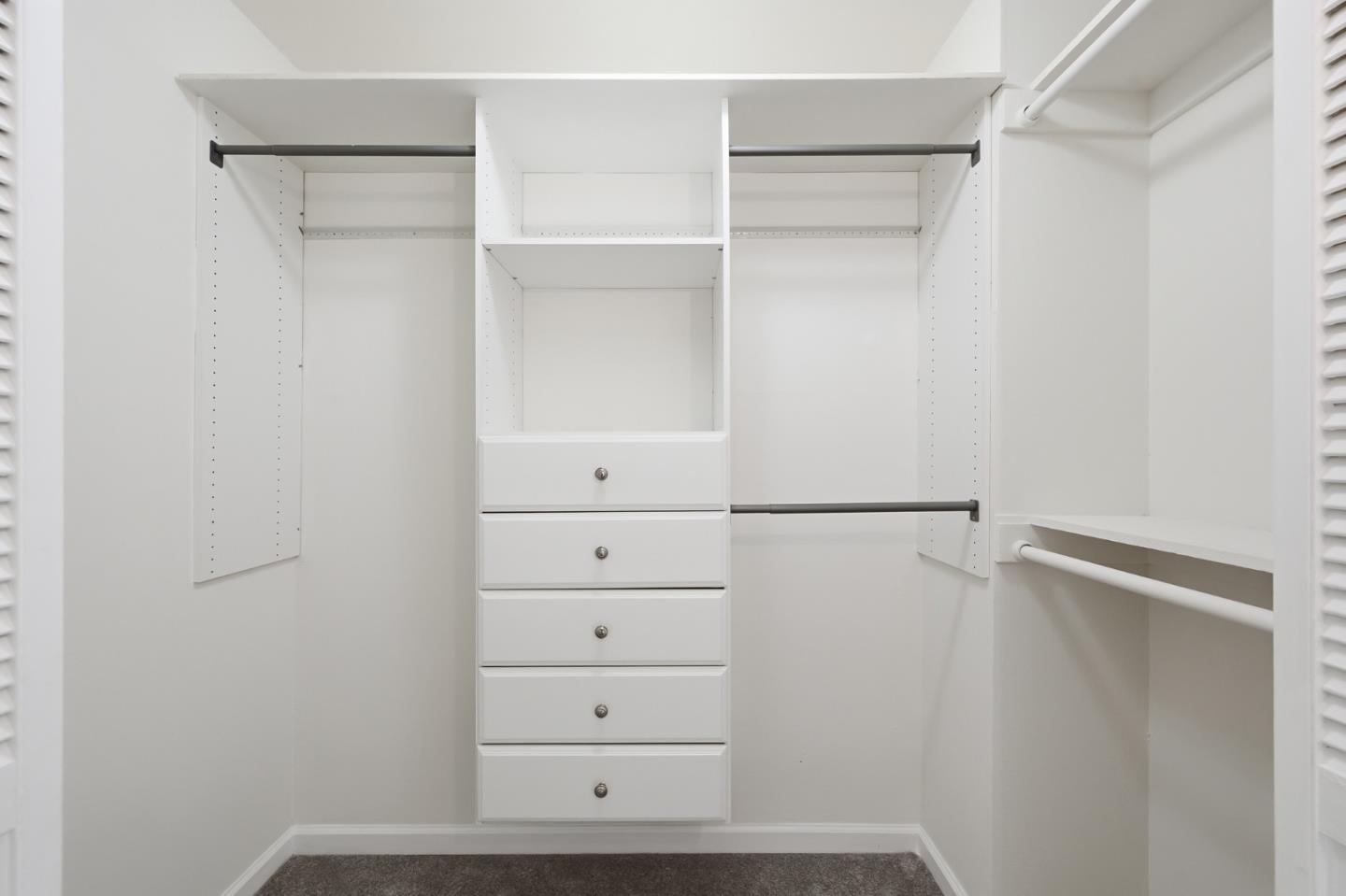 Walk-in closet with white built-in shelves, hanging rods, and five central drawers.