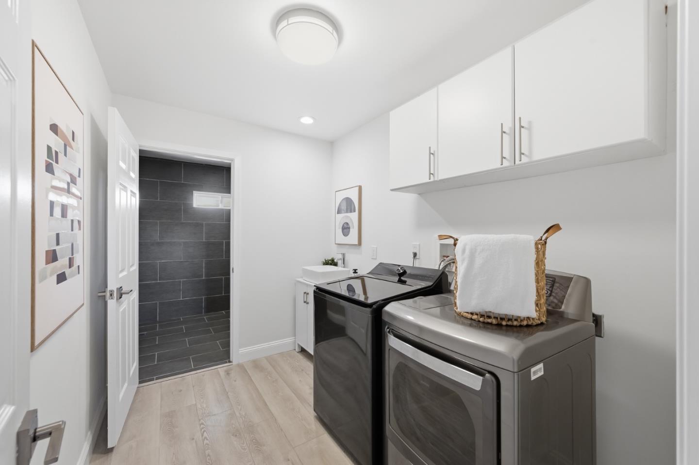 Laundry room with front-loading washer and dryer beneath white cabinets, opening to a dark-tiled bathroom.