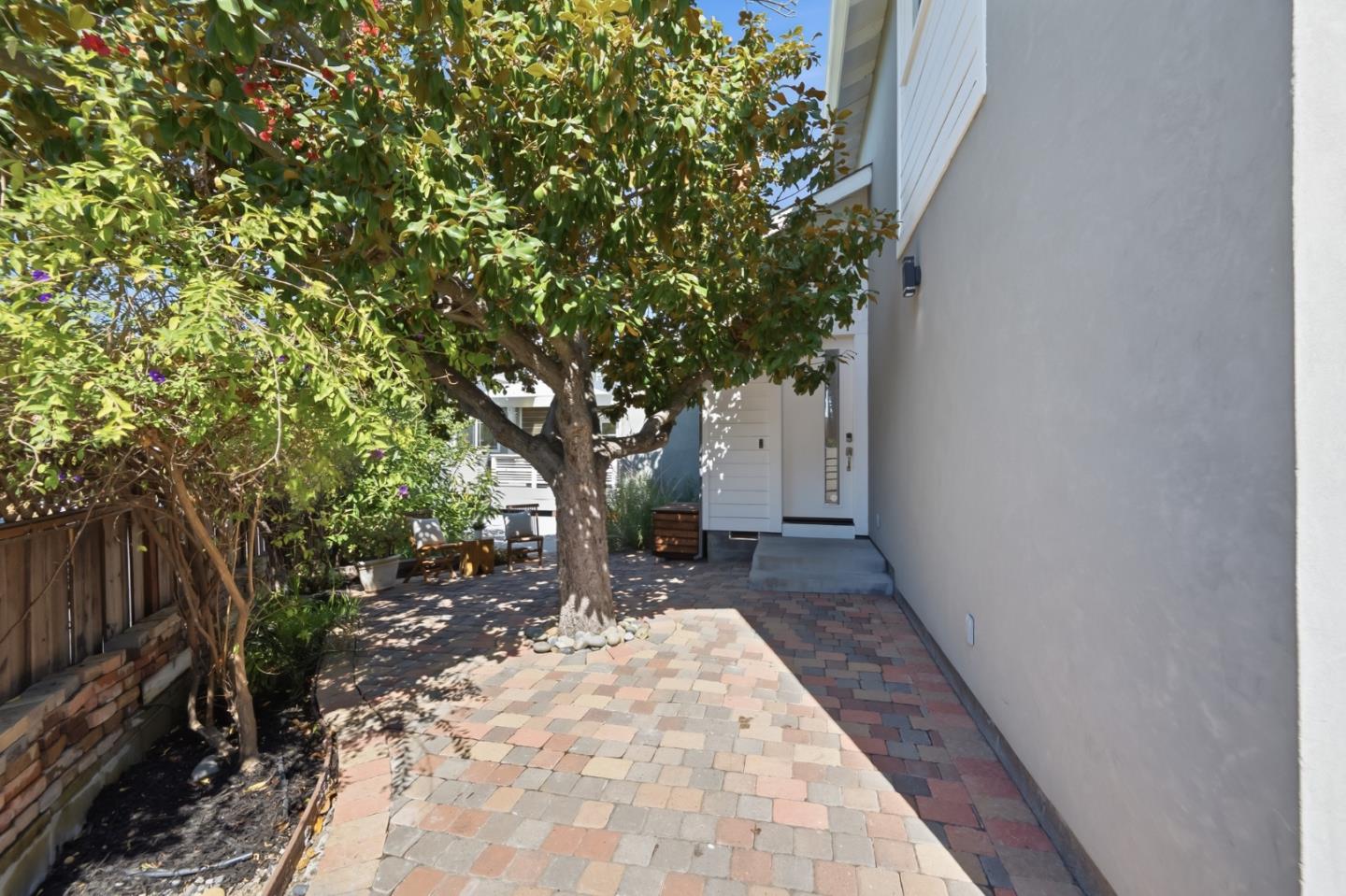 Brick-paved entry courtyard with a mature tree and seating beside the white front door.