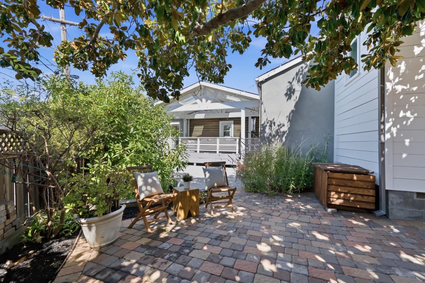 Stone-paver patio with wooden lounge chairs, leafy planters, and a shaded house facade.