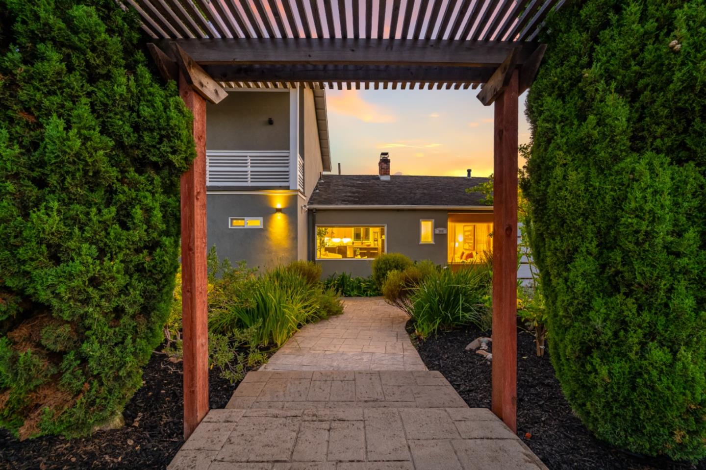 Gray two-story home glowing at dusk beyond a pergola-framed path and landscaped hedges.
