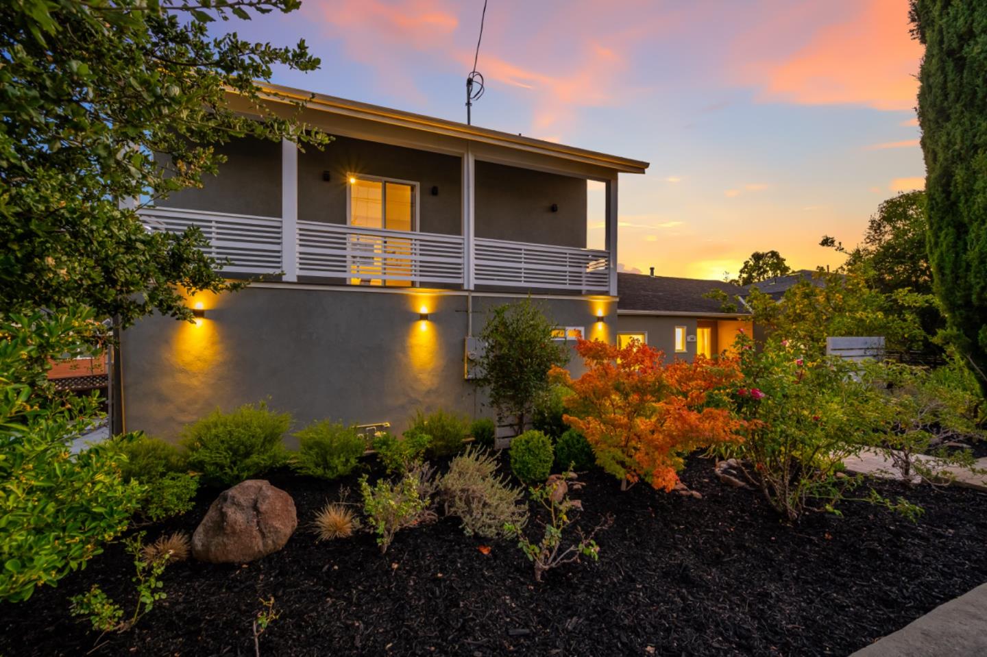 Two-story gray home with a white-railed balcony and landscaped garden at dusk.
