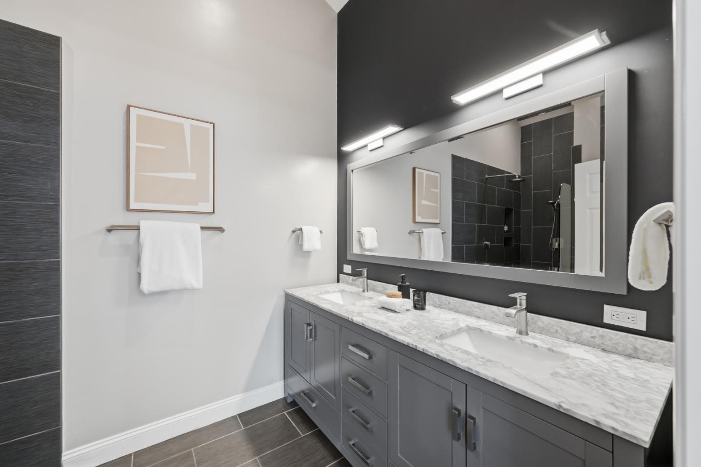 Modern bathroom with a gray double vanity, marble-look countertop, illuminated mirror, and dark tiled shower.