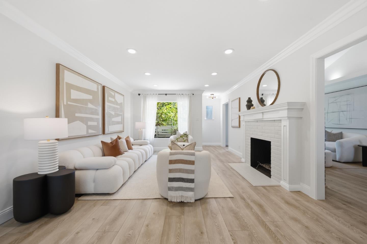 Staged living room with a white sofa, brick fireplace, and window overlooking greenery.