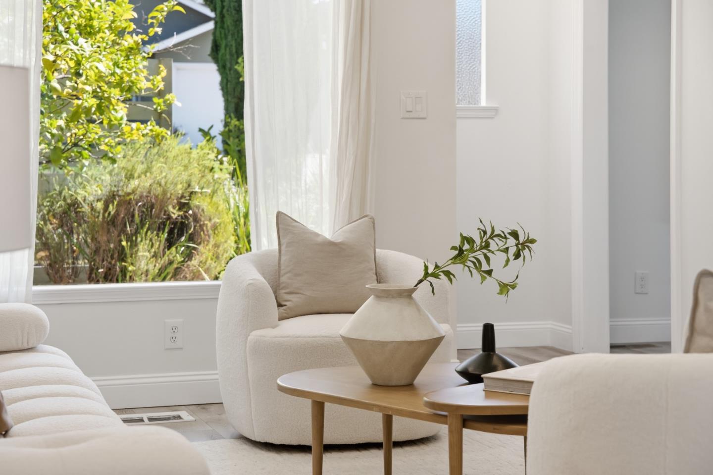 Bright living room with white upholstered chairs, a wood coffee table, and a garden view through a large window.