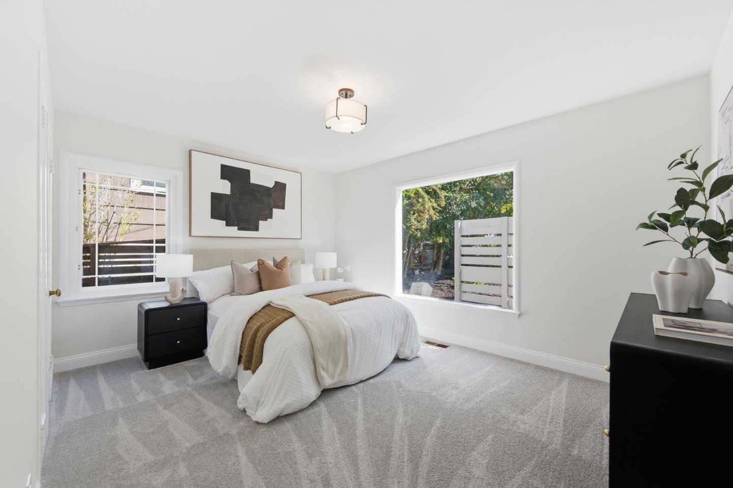 Staged bedroom with a white bed, tan throw, abstract wall art, and windows overlooking greenery.