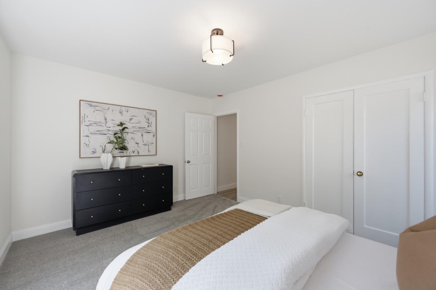 Bedroom with a white bed, tan knitted runner, black dresser, and framed abstract art.